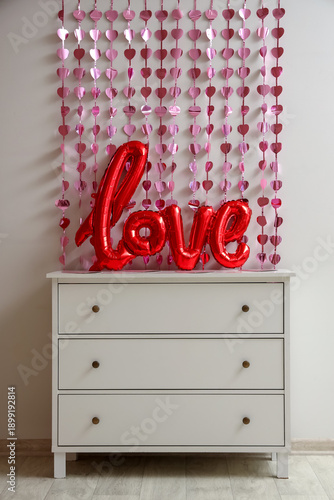 Chest of drawers with word LOVE made from balloons and pink hearts garland near white wall. Valentine's Day celebration
