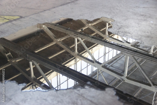 A puddle of fuel and water on a concrete floor with reflections of the ceiling and window structures inside the building