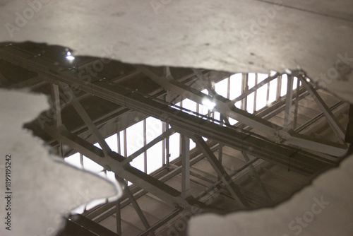 A puddle of fuel and water on a concrete floor with reflections of the ceiling and window structures inside the building