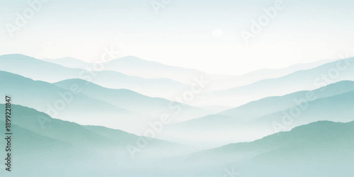 A misty mountain landscape at dawn features snow-capped peaks rising above a foggy valley under a blue horizon sky as the morning sun breaks through the clouds and haze