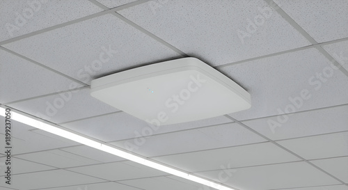 White ceiling-mounted device in a modern office setting. Square, flat device mounted on a light gray tile ceiling.