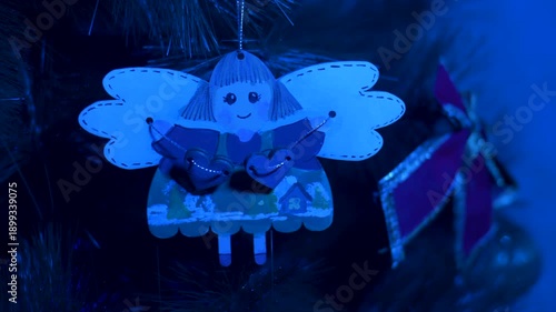 A warm, atmospheric shot of a Christmas angel ornament gently hanging on a holiday tree. Soft purple-blue lighting and festive lights create a cozy, magical mood. 
