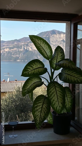 Cozy vintage Italian-style room with piano, green plant on the window, view of Lake Garda and the Alps outside, warm atmospheric interior scene.