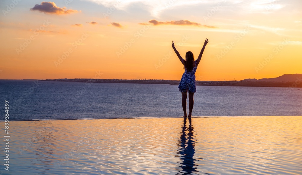 Fototapeta premium A girl stands on the edge of the pool against the background of a sea sunset
