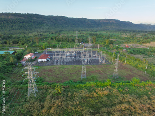 Distant aerial view of electrical substation and power pylons surrounded by green fields and forest.