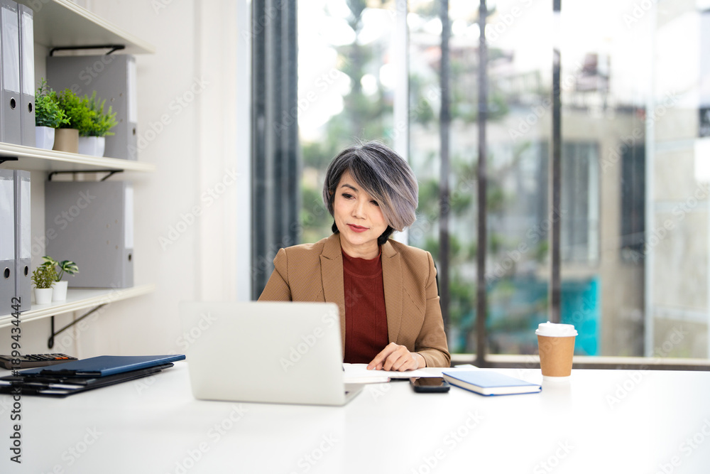 Fototapeta premium Confident mature woman, writer using laptop computer, taking notes, working project in modern office. financier working at workplace. Successful business