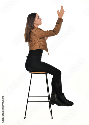 full length portrait of pretty brunette female model wearing basic modern  black clothing with brown leather jacket. sitting pose on chair, isolated figure on white studio background.