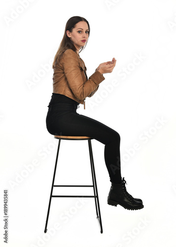 full length portrait of pretty brunette female model wearing basic modern  black clothing with brown leather jacket. sitting pose on chair, isolated figure on white studio background.
