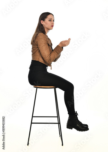 full length portrait of pretty brunette female model wearing basic modern  black clothing with brown leather jacket. sitting pose on chair, isolated figure on white studio background.