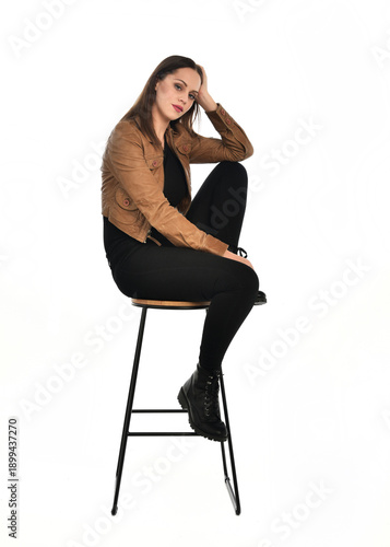 full length portrait of pretty brunette female model wearing basic modern  black clothing with brown leather jacket. sitting pose on chair, isolated figure on white studio background.
