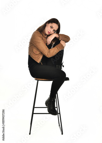 full length portrait of pretty brunette female model wearing basic modern  black clothing with brown leather jacket. sitting pose on chair, isolated figure on white studio background.
