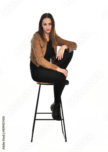 full length portrait of pretty brunette female model wearing basic modern  black clothing with brown leather jacket. sitting pose on chair, isolated figure on white studio background.
