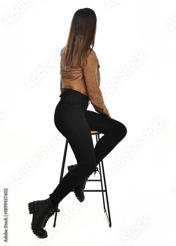 full length portrait of pretty brunette female model wearing basic modern  black clothing with brown leather jacket. sitting pose on chair, isolated figure on white studio background.