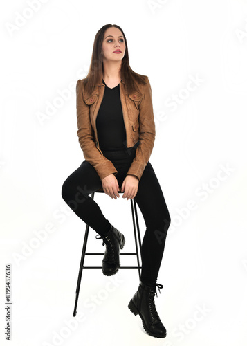 full length portrait of pretty brunette female model wearing basic modern  black clothing with brown leather jacket. sitting pose on chair, isolated figure on white studio background.

