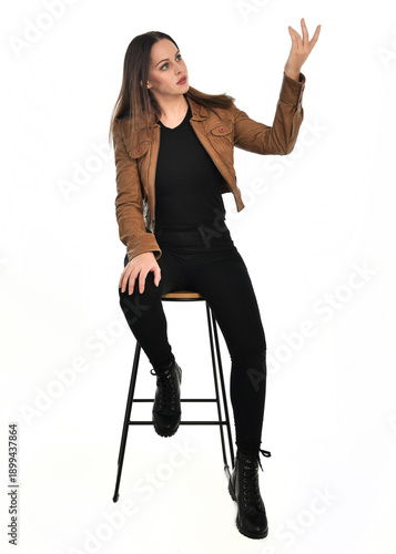 full length portrait of pretty brunette female model wearing basic modern  black clothing with brown leather jacket. sitting pose on chair, isolated figure on white studio background.
