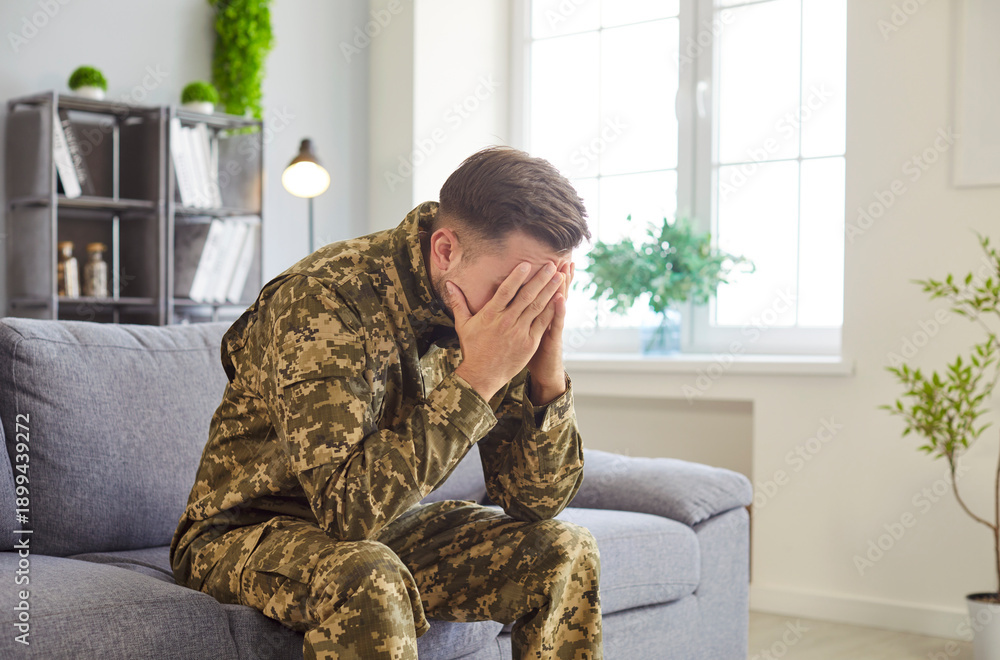 Fototapeta premium Man in military camouflage uniform sits on couch expressing deep sadness or emotional stress, suffers from post-traumatic stress disorder, need psychological aid. Mental health, counselling