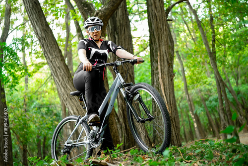 Woman athlete riding mountain bike through dense green forest trees. Extreme cyclocross activity for building physical strength and enjoying wilderness adventure.