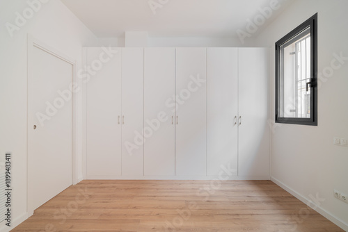 Wallpaper Mural A minimalist interior of a room with white walls and a wooden floor. A large white wardrobe occupies one wall. A window allows natural light to enter. Torontodigital.ca