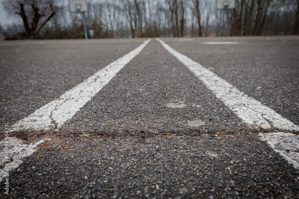 Obraz premium Faded white lines stretch across a weathered asphalt court, leading towards distant, blurry basketball hoops amidst bare trees. The scene evokes a sense of quiet abandonment and past games.