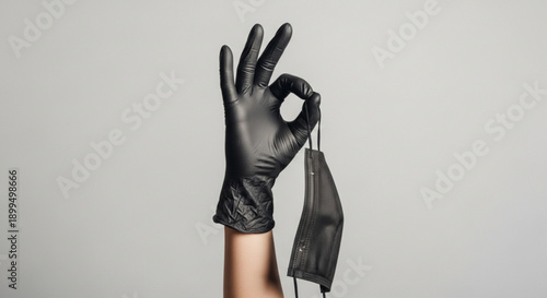 Hand in black glove holding face mask with okay sign on gray background symbolizing safety and protection