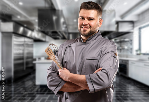 Smiling male chef in gray uniform holding whisk and wooden spatula in professional kitchen