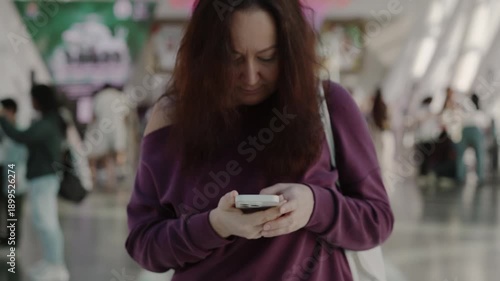 Middle aged caucasian woman standing indoors holding smartphone with serious expression in modern public interior, symbolizing technology use and everyday communication.
