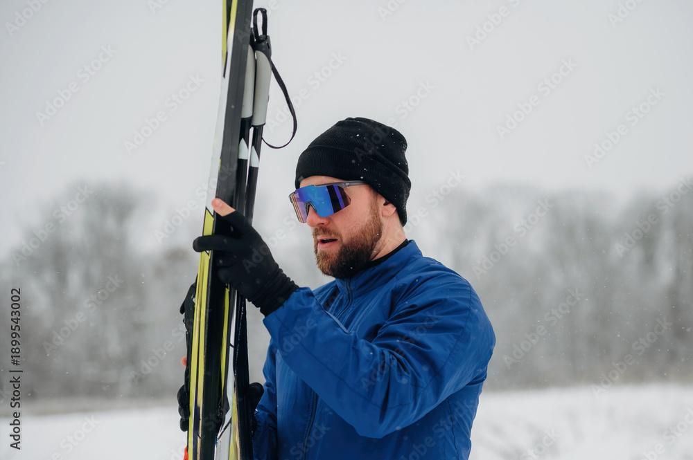 Naklejka premium Male skier in blue jacket and black beanie adjusts ski poles while standing in snowy landscape with blurred trees in the background