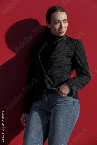 Woman posing against a red wall while wearing a black blazer and blue jeans