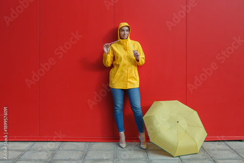 Woman in yellow raincoat holding a zipper standing by red wall with umbrella on ground