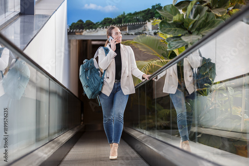 Woman using phone while walking along a glass corridor with backpack in modern setting