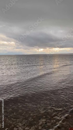 The seashore in cloudy weather, calm water, calm water in the evening. Sun rays behind the clouds.