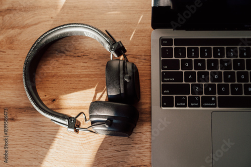 Top view of black wireless headphones lying on a wooden desk next to a silver laptop. Warm natural sunlight creates soft shadows on a modern, minimalist home office workspace.