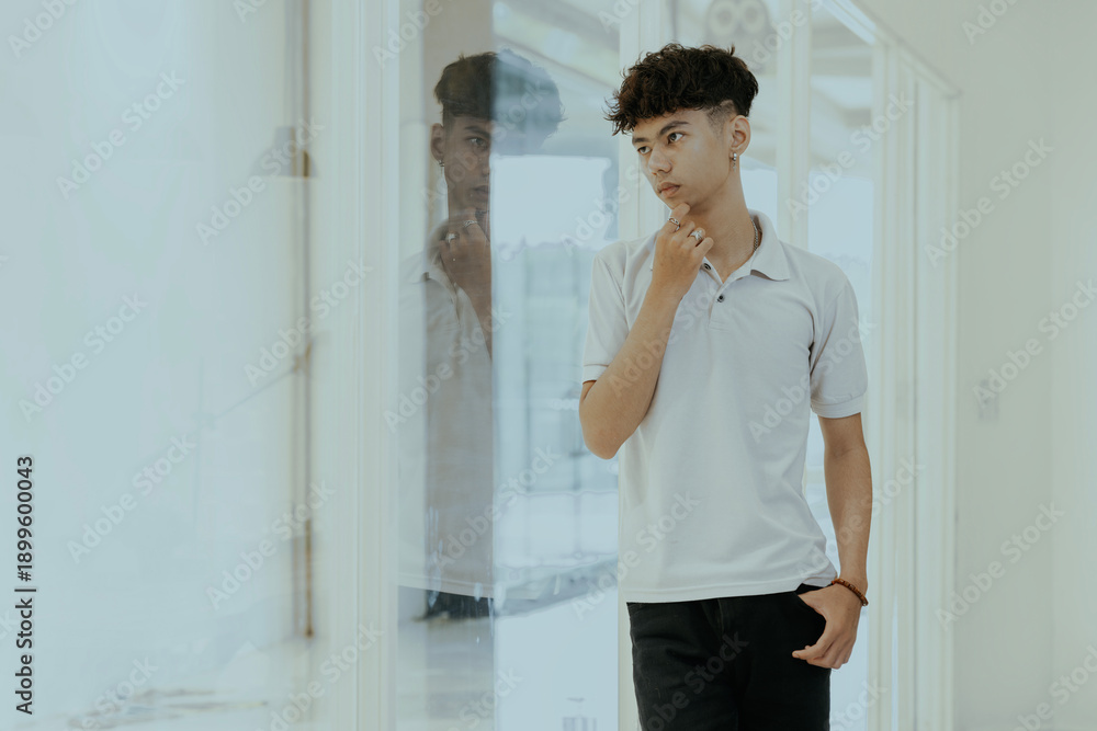 Obraz premium Handsome young asian man standing by the window, thoughtfully looking out with his hand resting on his chin. He appears pensive and deep in thought.