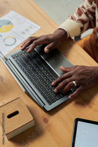 Black man typing on laptop at desk, hands with wedding ring, working on project with financial charts and tablet nearby, adult engaged in professional activity