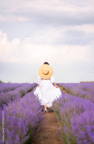 a beautiful woman in a long white dress and a large braided hat in a lavender field. View from the back. In the hands of a basket with flowers