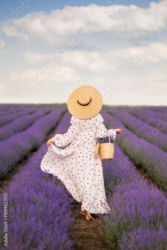 a beautiful woman in a long white dress and a large braided hat in a lavender field. View from the back. In the hands of a basket with flowers
