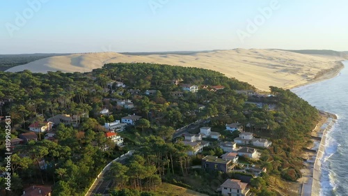 Panorama of the French Atlantic coast near Arcachon. In the foreground, a wooded coastline with exclusive villas stretches out, while the massive silhouette of the Dune du Pilat rises on the horizon.