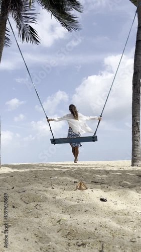 A woman in a white top and blue patterned skirt sits on a wooden swing suspended between two palm trees on a sandy beach. 