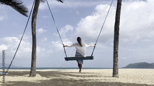 A woman in a white top and blue patterned skirt sits on a wooden swing suspended between two palm trees on a sandy beach. 
