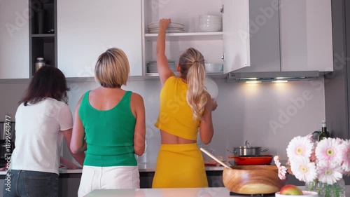Female friends in modern kitchen holding plates and talking while preparing for meal, surrounded by kitchenware, fresh fruit, and flowers on counter, showing casual indoor domestic activity