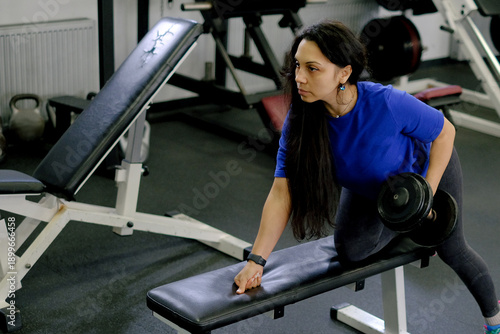 Woman doing dumbbell row supported on bench in gym