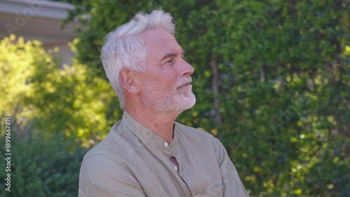 Confident senior businessman with white hair and beard standing with arms crossed near greenery and office building on sunny day