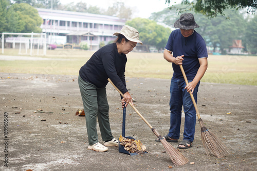Two Asian adults cleaning public area by sweeping dry leaves together. Concept, Community service, public cleanliness, environmental care, teamwork, volunteer activity and responsible lifestyle. 