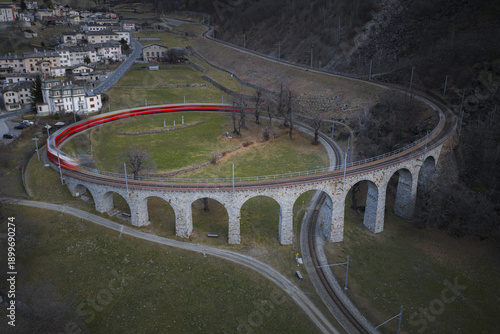 Aerial view of the Brusio spiral viaduct curving gracefully through the landscape with a red train leaving a light trail, Brusio, Grisons, Switzerland.