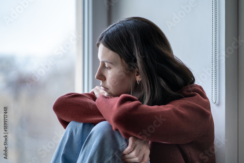Quadro su tela Sad reflective teenage girl sits hugging knees looking to window