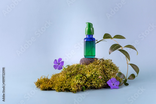 bottle of cosmetics stands on moss next to periwinkle flowers on a blue background