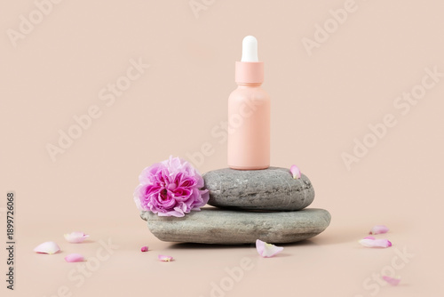 bottle with an essential oil pipette on a stand made of natural stones, petals and a wild rose flower on a beige background