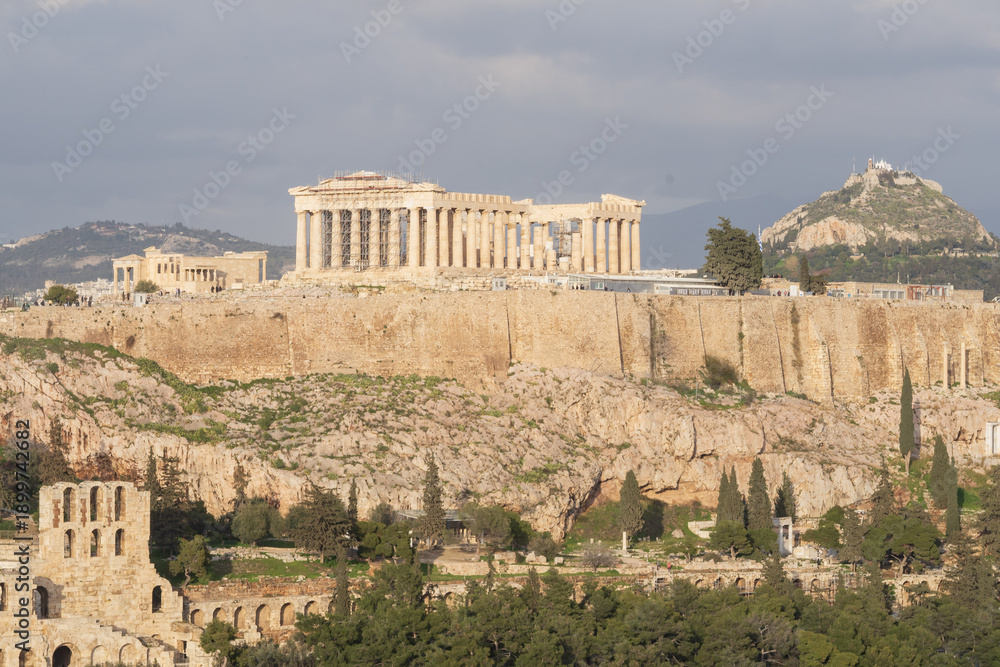 Obraz premium The Acropolis of Athens with the Parthenon and Odeon of Herodes Atticus, seen from Filopappou Hill at sunset. Golden hour light, ancient ruins, rocky landscape and historic Greek landmarks.