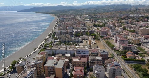 A wide aerial panorama of a coastal town, featuring a long stretch of pebble beach bordering the blue sea, with dense residential buildings. It's Catanzaro Lido seafront, in Calabria, Italy.
