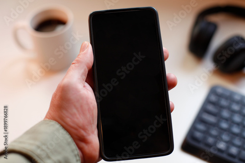 Hand Holding Black Smartphone With Blank Screen In Modern Workspace Setup